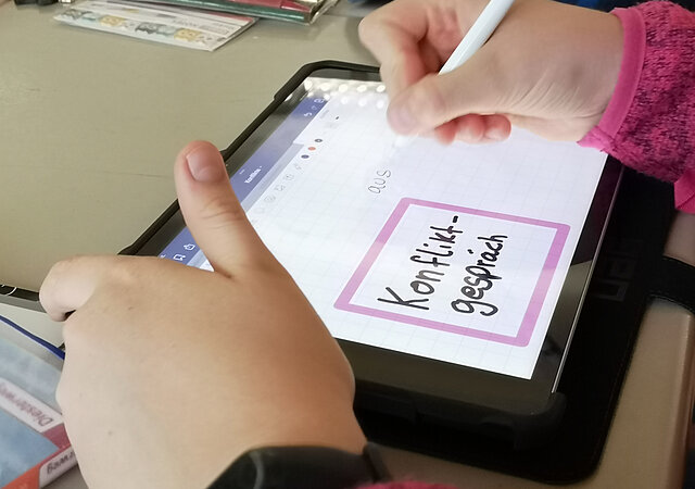 Nahaufnahme eines iPads in den Händen eines Schulkindes. Auf dem Screen steht rosa umrandet das Wort "Konfliktgespräch".