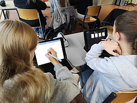 Zwei Mädchen arbeiten im Unterricht an ihren iPads. Aufnahme von hinten über ihre Schultern. 