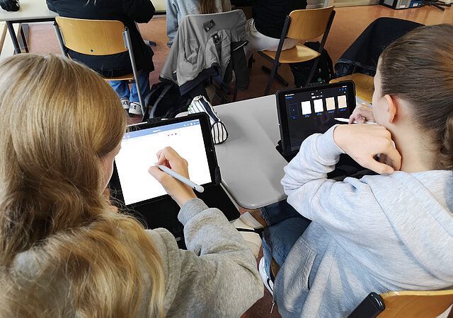 Zwei Mädchen arbeiten im Unterricht an ihren iPads. Aufnahme von hinten über ihre Schultern. 
